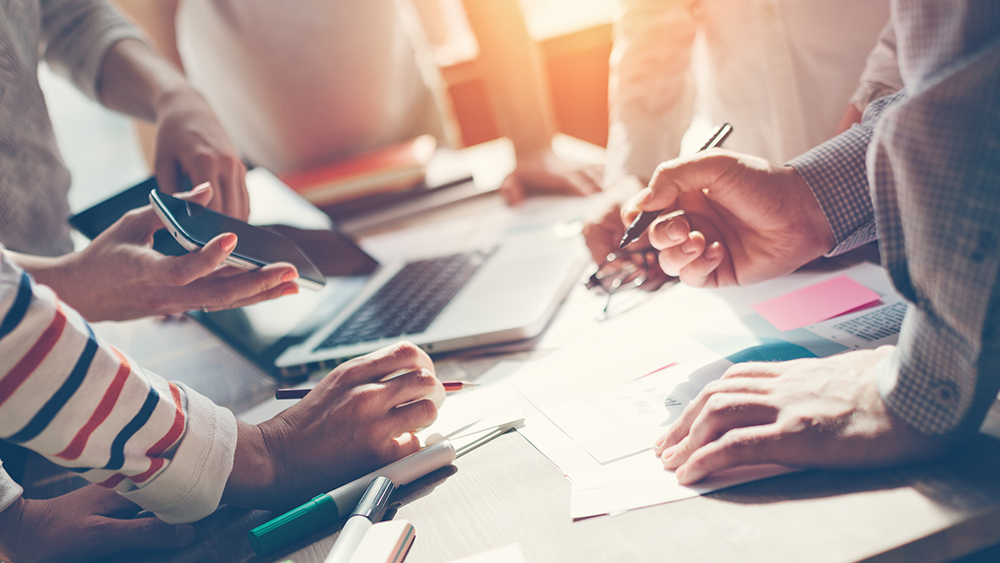 Kokouspöytä, jossa papereita ja tietokone. Ihmisten käsiä pöydän ympärillä..